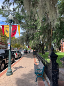A sidewalk in New Bern NC has a lamppost with New Bern flags on the left, and a tree with Spanish moss and a bench and a decorative black iron fence on the right. New Bern is located on the "Inner Banks" of the Crystal Coast.